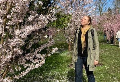 Nainen hymyilee kukkivan pensaan vierellä auringonpaisteessa